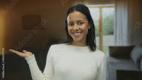 Photos Woman smiling indoors, extending hand in a cozy room, wearing white sweater, suggesting hospitality with a joyful expression, creating a warm and inviting atmosphere