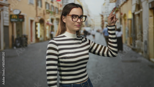 Foto Woman wearing striped sweater and tortoiseshell glasses pointing index finger on a narrow cobblestone street with blurred pedestrians and storefronts; quiet confidence