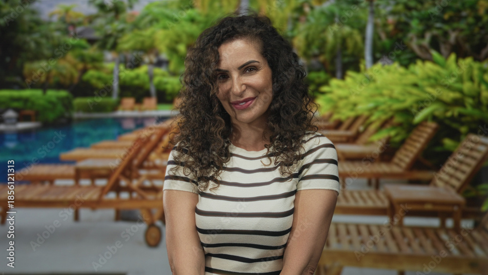 Naklejka premium Woman smiles with bare arms and curly hair while tilting head at resort poolside under bright sunlight; serenity.