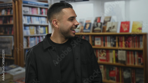 Slika na platnu Young man wearing black jacket smiles and turns head near bookshelf while speaking in library; curiosity exploration