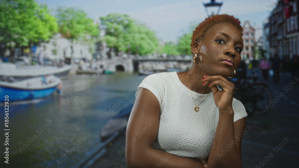 Fototapeta premium Woman in white top with hand on chin and crossed arms on sunlit street by canal boats; introspection.