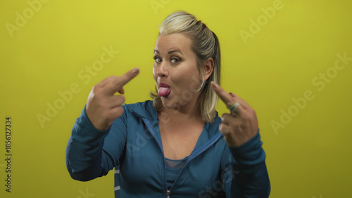 Obraz na plátně Woman showing middle finger gesture in yellow studio wearing blue zippered jacket and smiling broadly; defiance
