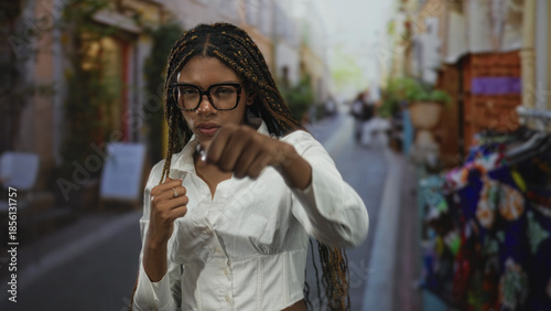 Fotografie Woman holding fists up in a guarded boxing stance on a bustling city street with braided hair, glasses and white shirt visible; determination