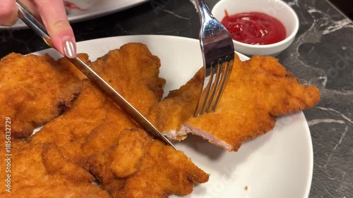 Girl eats with fork and knife appetizing chicken schnitzel and dips it in ketchup red sauce traditional Austrian food restaurant white plate meat in Klaar
