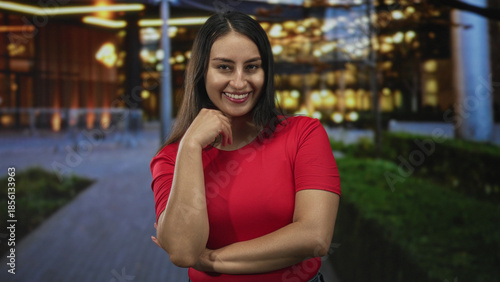 Young hispanic woman smiling with hand on chin and crossed arms in front of lit building facade at night; confidence warmth friendliness. Free Download Young hispanic woman smiling with hand on chin and crossed arms in front of lit building facade at night; confidence warmth friendliness. - GFXLoad