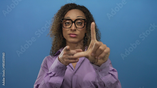 Photos Hispanic transgender woman wearing purple blouse forms l shape gesture with raised hand in blue studio; mockery