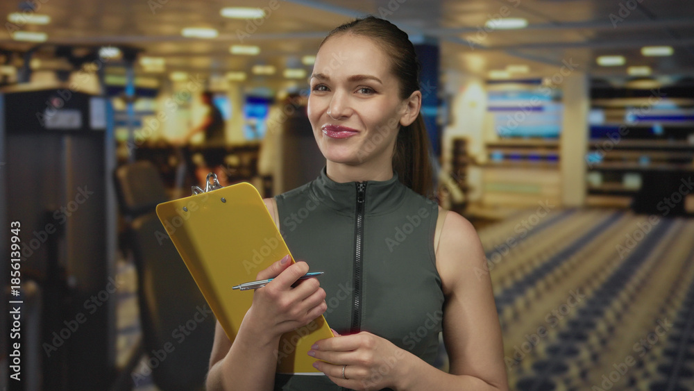 Fototapeta premium Woman with clipboard and pen gestures while discussing workout routine in a gym building; motivational support.