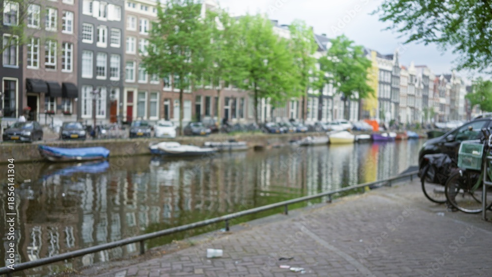 Obraz premium Blurred view of amsterdam's iconic canals with classic buildings and moored boats casting reflections on water on a calm day.