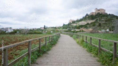 Photos Blurred view of a wooden path leading to a distant hillside castle with out of focus lush greenery under a cloudy sky, emphasizing a tranquil arta tourism scene in mallorca