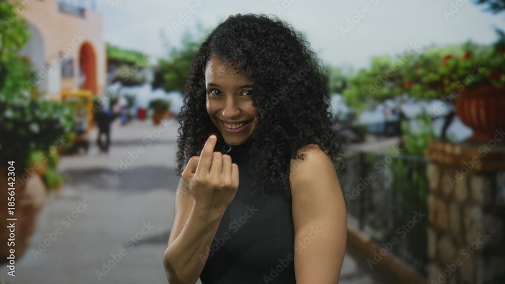 Fototapeta premium Smiling young hispanic woman beckons with finger on a sunlit outdoor street; playfulness invitation.
