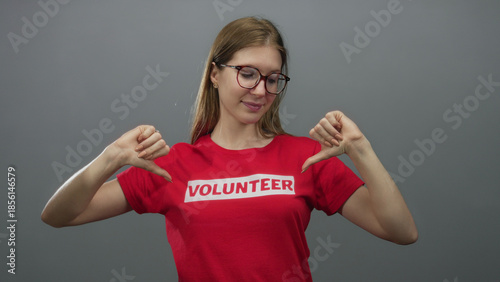 Tableau sur toile Woman in red volunteer shirt standing confidently with thumbs pointing to chest in a grey studio; pride