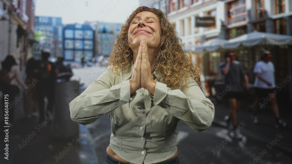 Fototapeta premium Woman middle age hispanic praying with hands pressed together, eyes closed and uplifted face, striped shirt visible while standing in a busy street cafe among storefronts and pedestrians; gratitude.