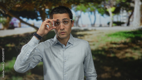 Young hispanic man adjusting glasses in a sunny park with trees and grass surrounding him, conveying a relaxed outdoor atmosphere.