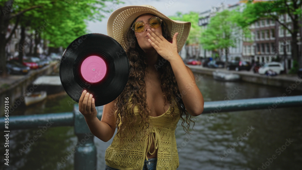 Fototapeta premium Hispanic woman holding vinyl record with hand, smiling and waving on a street bridge while wearing sunhat and sunglasses; joyful carefree summer.
