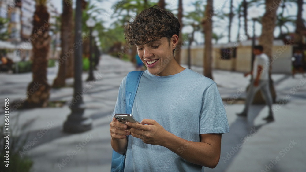 Fototapeta premium Teen boy smiling at smartphone, holding phone in hands with backpack strap over shoulder on a palm lined street; carefree joy.