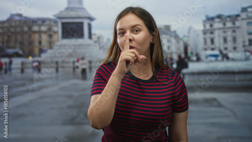 Fényképezés Woman with finger to lips and raised bare hand by monument on street wearing red navy striped tshirt and side glance; silence secrecy