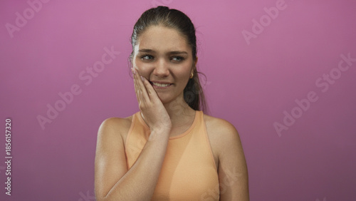 Bilde på lerret Teen girl wearing orange tank top holds cheek with hand and winces in pink studio; toothache severe pain