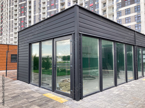 Modern, black prefabricated room on balcony, contrasting floor tiles, urban residential setting with multi-story building and several balconies, large windows, natural light, realistic style