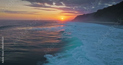 Golden sunset over Indian Ocean coastline, bright colorful clouds sky reflects on vast water surface, powerful waves break tropical Bali island coast with lush green cliffs in background. Aerial shot