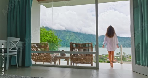 Indonesia, Bali, Batur: Woman in white shirt walk barefoot in modern hotel room to outdoor terrace with rattan chairs and table, enjoy beautiful mountain and lake landscape. Travel, holiday, vacation
