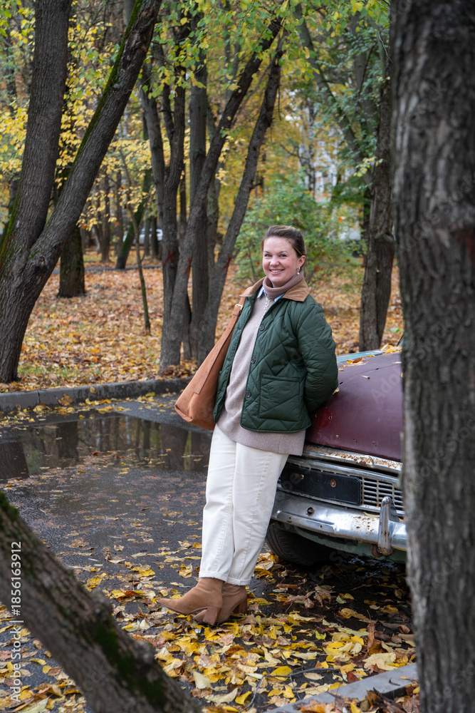 Fototapeta premium Smiling woman stands by a vintage turquoise car in a quiet autumn courtyard, fallen leaves, muted green and yellow tones, calm urban atmosphere