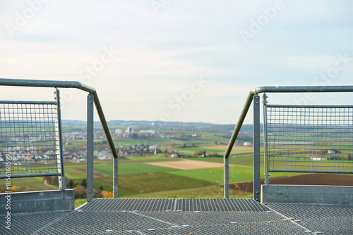 Metal platform overlooking farmland, town and hills under clear daylight sky. Railings and fences made of galvanized steel. Observation platform overlooking rural landscape.