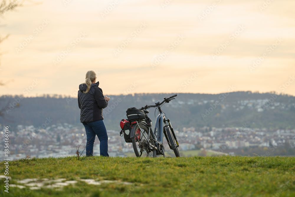 Obraz premium Cyclist on hilltop overlooking city. Person with bicycle on hilltop with gear, overlooking city, forest and evening sky.