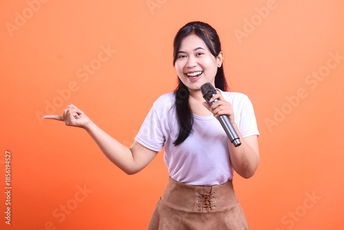 A smiling woman in a white shirt and brown skirt holding a microphone while pointing to the side, looking happy and present. isolated on orange background