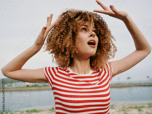 Fotografi A joyful woman with curly hair raises her arms in a candid outdoors portrait, wearing a red and white striped shirt, beside a calm water body and distant shoreline