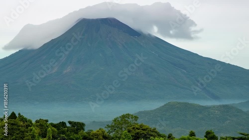 A large, verdant volcano looms, capped by clouds, above a forested landscape