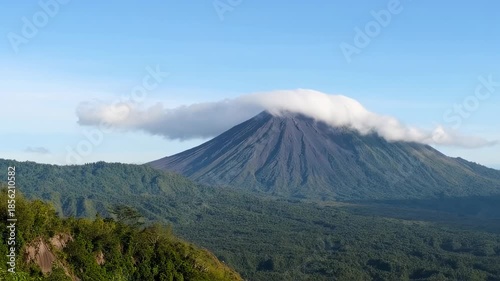 A volcanic mountain, partially veiled by clouds, rises above lush, green hills and clear sky