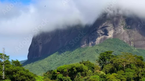 Majestic mountains shrouded in clouds rise above lush green forest in a sunlit landscape