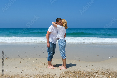 Couple standing barefoot on the beach, embracing near the waves with clear sky and casual outfits.
