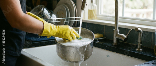 Person washing pot in kitchen sink with soap and water using yellow gloves