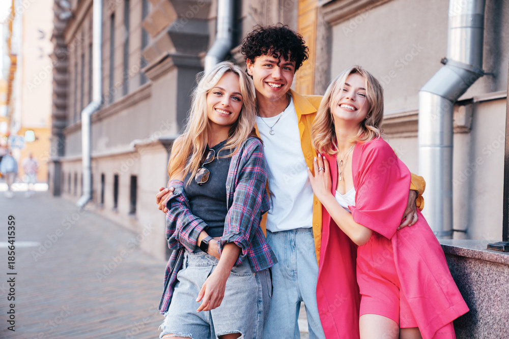 Fototapeta premium Group of young three stylish friends posing in the street. Fashion man and two cute female dressed in casual summer clothes. Smiling models having fun. Cheerful women and guy outdoors