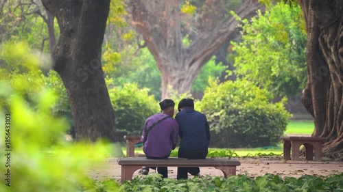 Young indian men sitting together on a park bench leaning on each other enjoying a cozy peaceful morning showing the various forms love and romance can take in the middle of trees and park