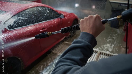 A young driver methodically applies thick detergent foam to his red sports car using a high-pressure hose gun at a modern self-service car wash station.