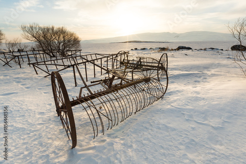 Vintage hay rake abandoned in snow at Lake Cildir, Kars, Turkey. Rustic agricultural machinery on frozen shore during winter golden hour with mountains.