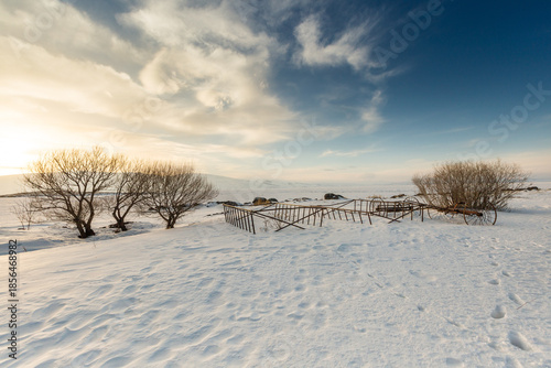 Vintage hay rake abandoned in snow at Lake Cildir, Kars, Turkey. Rustic agricultural machinery on frozen shore during winter golden hour with mountains.