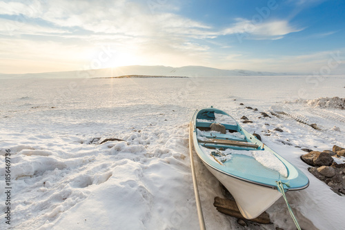 Blue fishing boat on frozen Lake Cildir at sunset, Arpacay, Kars, Turkey. Winter landscape with icy lake surface, snowy mountains and golden hour sunlight.