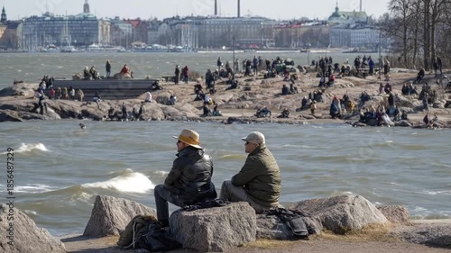 Two individuals sit on rugged rocks by a sprawling body of water, their backs to the viewer, as they observe the choppy waves and distant horizon. Further along the rocky shoreline, a diverse group of