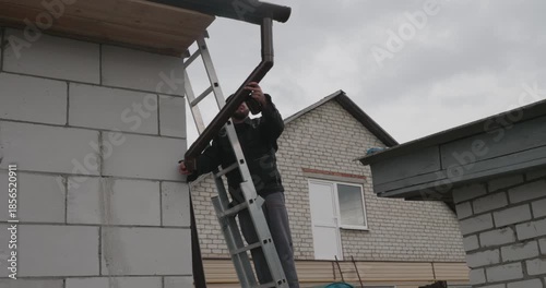 A Caucasian male contractor inspector inspects a drainpipe. A methodical worker checks the alignment of a drainpipe on a ladder next to a country brick house, measuring and adjusting a bracket.