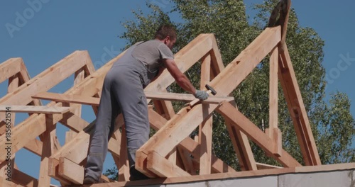 A construction worker uses a hammer to install wooden rafters and beams on the roof structure of a new home. The man secures wooden joists to the roof of the house. Roof installation.