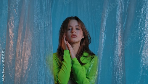Portrait of a young female model in a green top, posing against a blue background with transparent plastic curtains. Color beauty, fashion art.