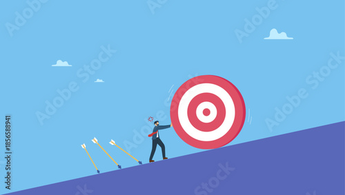 Businessman trying hard to push dartboard or arrow target up the hill. Missed target, failure or obstacle, difficulty in work that hard to achieve target or set too high or unrealistic goal.