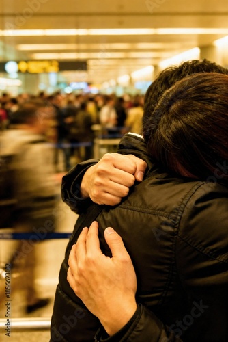 A tight shot of a powerful hug at an airport arrivals gate, a white-knuckled grip and buried head, with the crowd blurred into a motion background.
