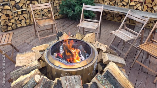 Outdoor Fire Pit Surrounded By Wooden Chairs And Stacked Firewood, Cozy backyard seating area with burning logs creating warm rustic atmosphere