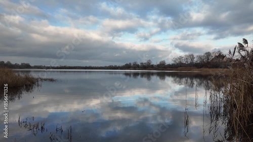Wide panoramic view of a calm river or lake with golden reeds, dramatic cloudy sky and soft reflections on water. Peaceful autumn wetland landscape, natural light, rural scenery.