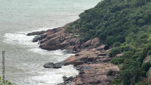 Waves strike rugged coastal rocks as powerful surf spreads white foam across the shoreline while a touch of greenery above the stones softens the dramatic look of the stormy water. Coastal force.