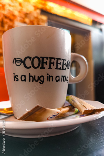 Close-up of a white coffee mug reading Coffee is a hug in a mug, resting on a saucer with discarded sugar packets in a cafe